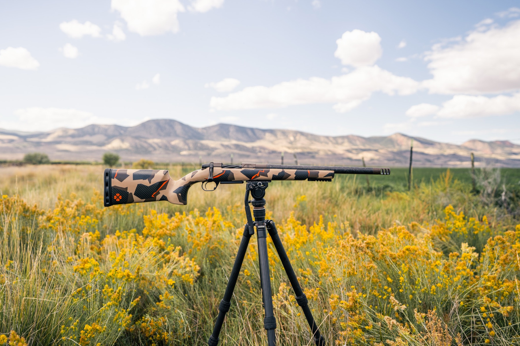 Rifle with a shard camo carbon stock and burnt bronze action, on a tripod in sagebrush below mountains